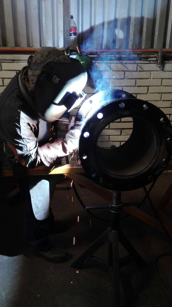 Soldador ejecutando soldadura SMAW con arco eléctrico visible sobre flange de gran diámetro